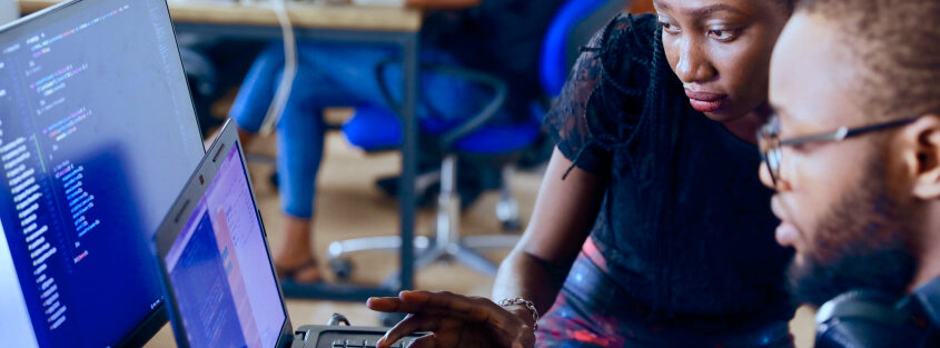 A man and woman analyzing code on a laptop and computer monitor together